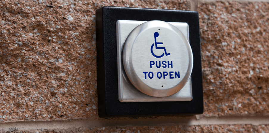 Symbolisch für digitale Barrierefreiheit: Schaltfläche an einer Wand mit einem Rollstuhl-Symbol und Aufschrift "Push to open"