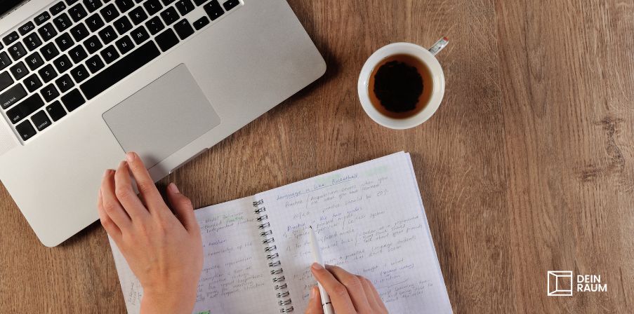 Auf einem Holztisch steht ein Laptop, ein Becher Kaffee und eine Person schreibt etwas in sein bereits volles Notizheft.