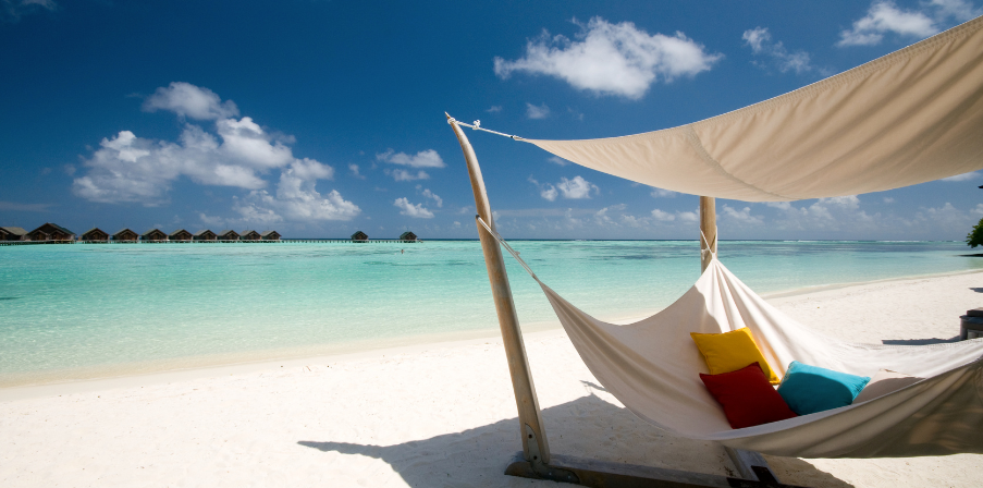 Traumhafter weißer Sandstrand, türkisblaues Meer und eine große Hängematte mit weißem Stoff und bunten Kissen - Sinnbild für Urlaub.