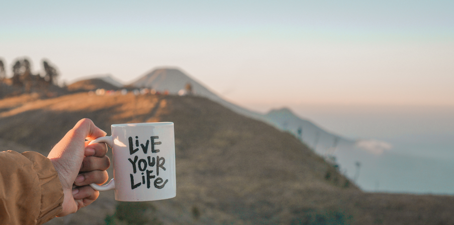 Sinnbild für Work-Life-Blending: Becher in der Hand mit der Aufschrift "Live your life" vor einer wunderschönen Bergkulisse zum Sonnenaufgang