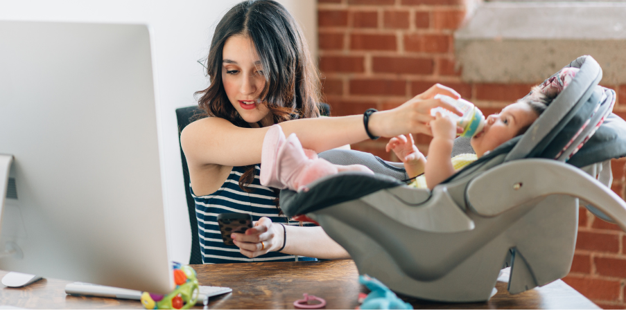 Sinnbild für Work-Life-Blending: eine junge Mutter vor einem Computerbildschirm mit einem Handy in der einen Hand und mit der anderen Hand ein Baby, in einer Babyschale ebenfalls auf dem Schreibtisch stehend, fütternd.