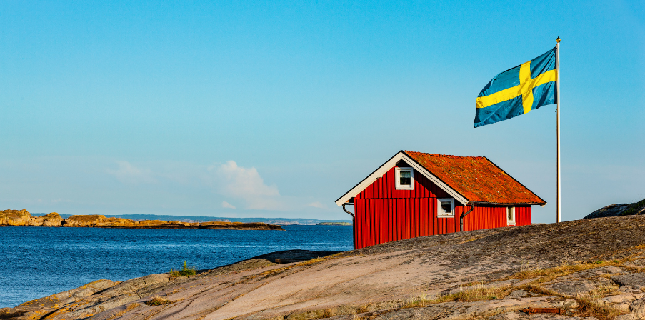 Sinnbild für Schweden als Vorreiter für das 30-Stunden-Woche-Modell: ein typisches rotes Holzhaus an einer Küste mit einer wehenden schwedischen Flagge  