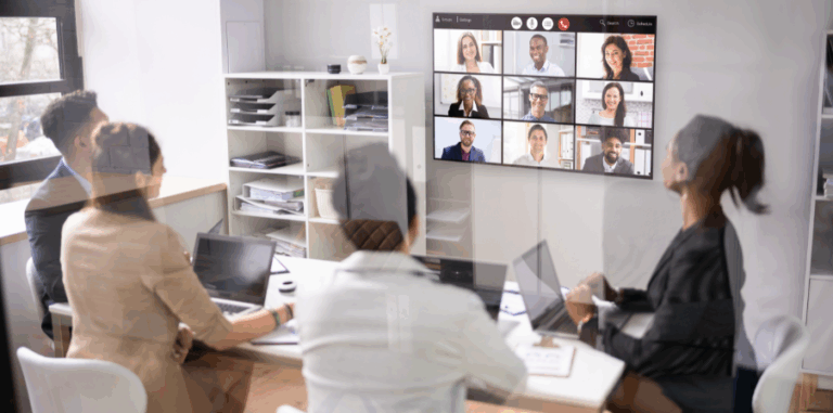 Symbolbild für hybrides Arbeiten. Vier Personen sitzen in einem Büro an einem Konferenztisch und nehmen an einer Videokonferenz mit neun weiteren Teilnehmenden auf einem großen Bildschirm teil.